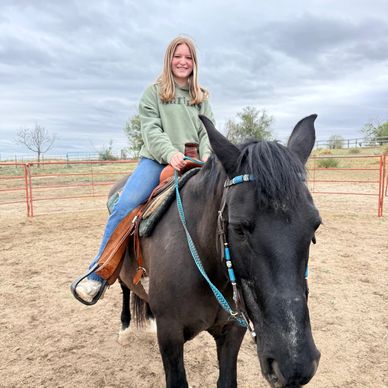 Young girl smiling while riding a black horse in an outdoor pen.
