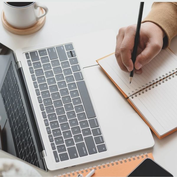 Person writing in a notebook beside a laptop and coffee cup.