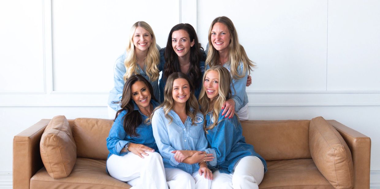 Six women smiling together on a brown sofa, wearing denim shirts and white pants.