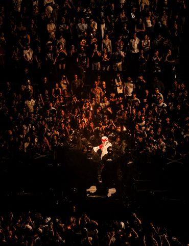 A performer in white illuminated amidst a dark crowd and security.