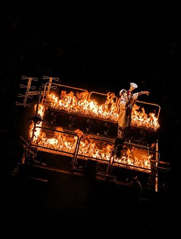 Performer stands with megaphone on flaming stage, surrounded by intense fire.