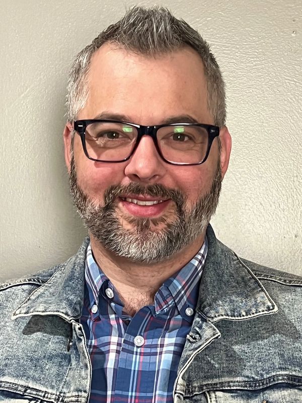 Man with glasses and beard wearing a denim jacket and plaid shirt.