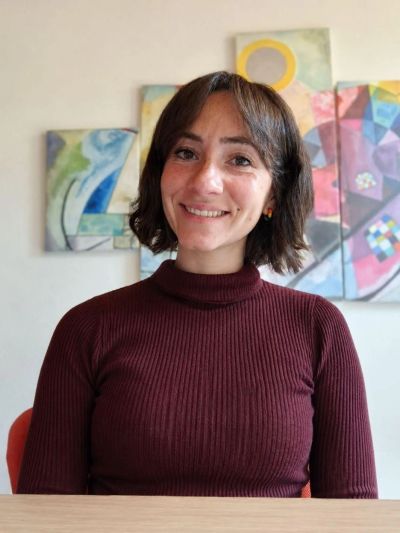 Smiling woman in a maroon turtleneck sitting indoors with colorful abstract paintings behind her.