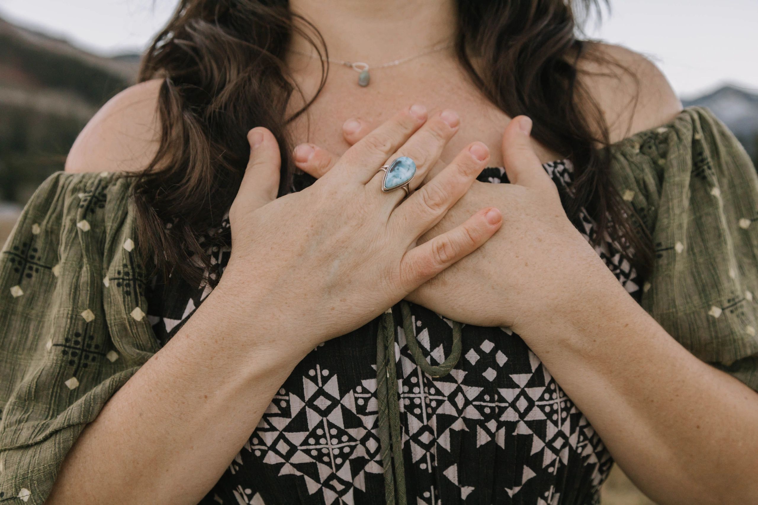 Woman with hands on chest wearing patterned dress and blue ring.