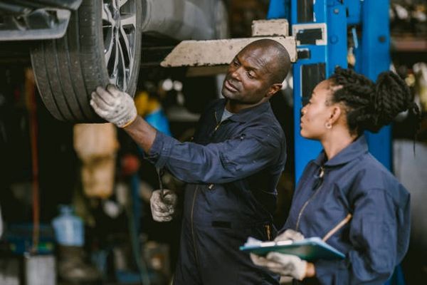 Two mechanics inspecting a car wheel in a garage, wearing navy blue coveralls.