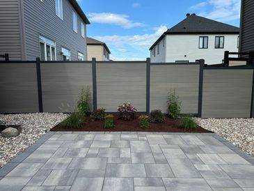 A modern backyard with a gray fence, paved patio, and small garden bed with plants.