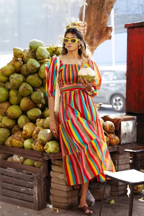 A woman in a colorful striped dress holds a coconut beside a pile of green coconuts.