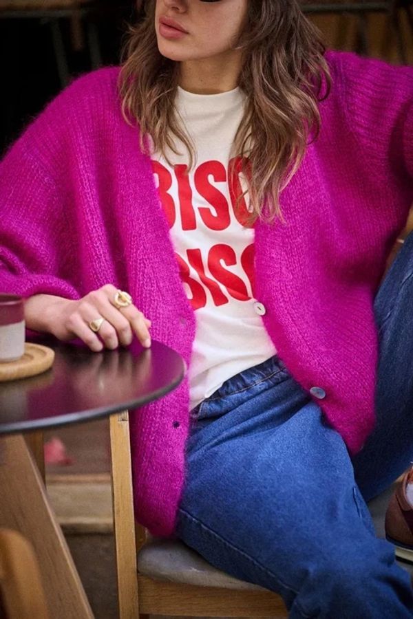Woman wearing a bright pink cardigan, graphic tee, and blue jeans sitting at a table.
