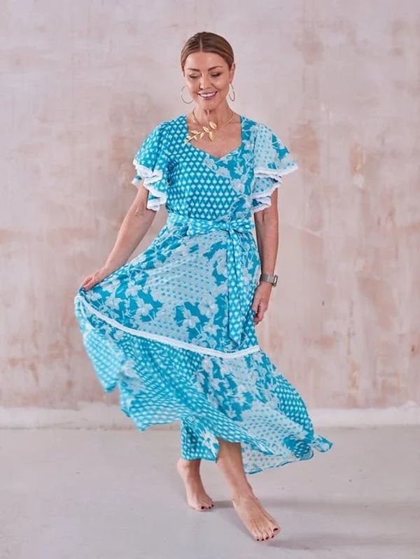 Woman in a flowing blue patterned dress smiling and posing barefoot.