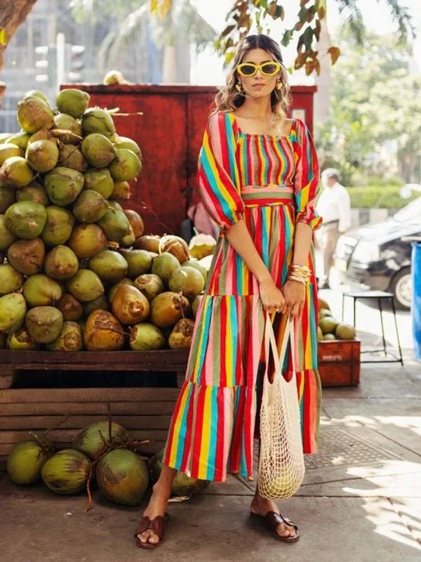 Woman in colorful striped dress with yellow sunglasses stands by coconuts outdoors.