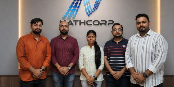 Five professionals stand in front of a Mathcorp logo posing for a group photo.