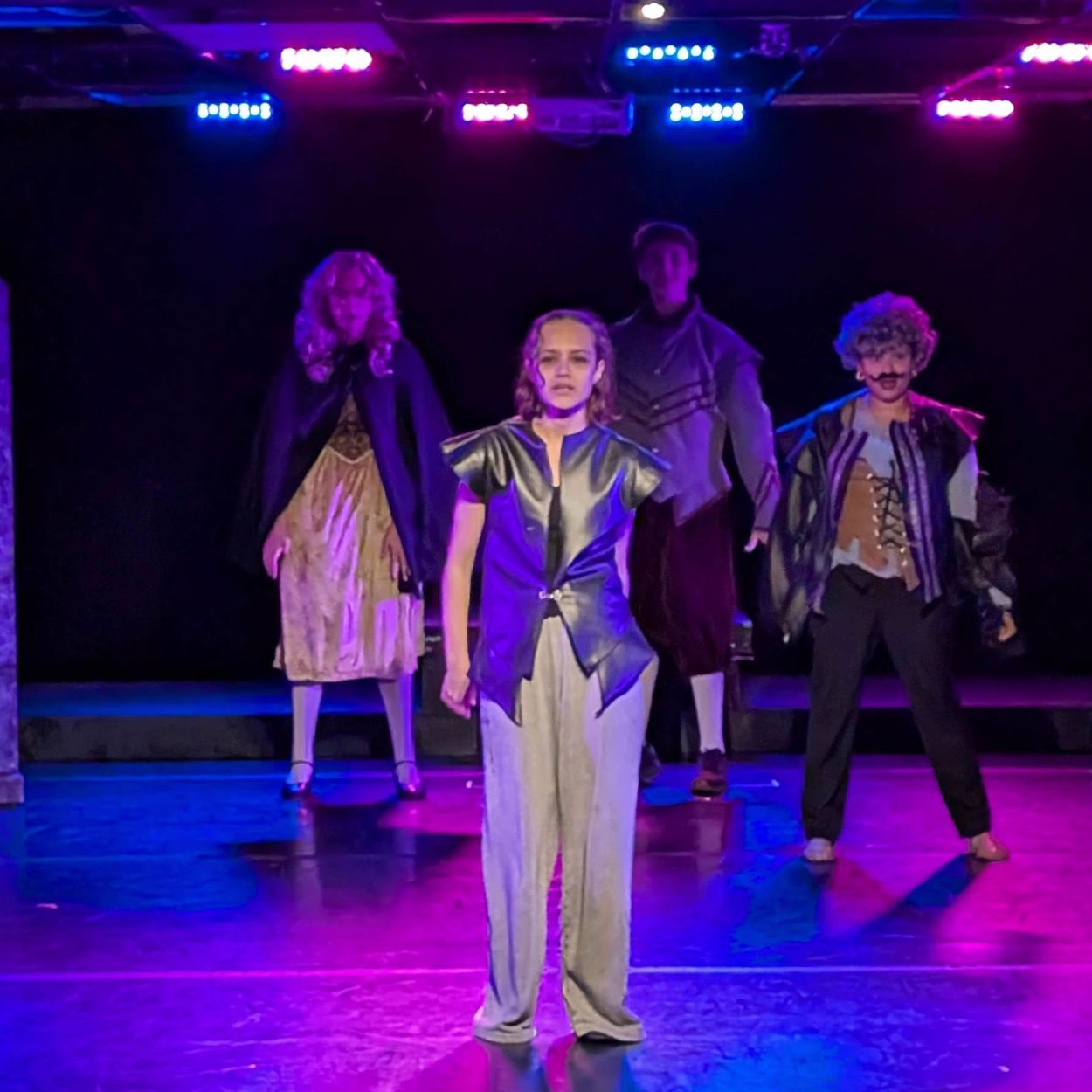 Actors perform on stage in dramatic costumes under colorful lights.