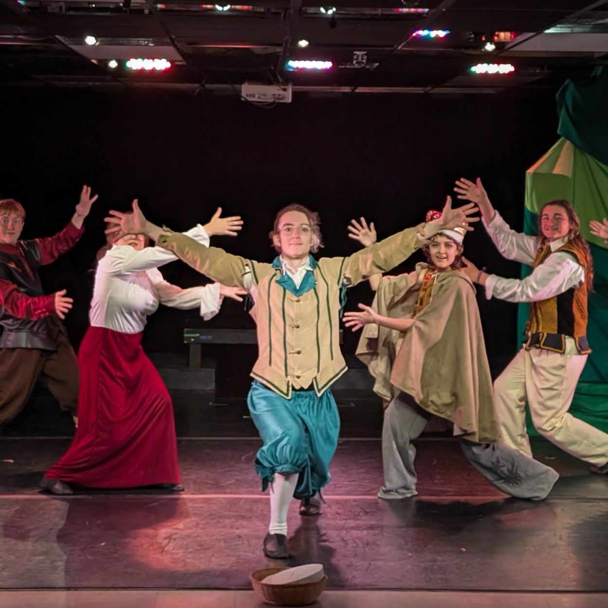 Actors in historical costumes perform on stage with open arms.