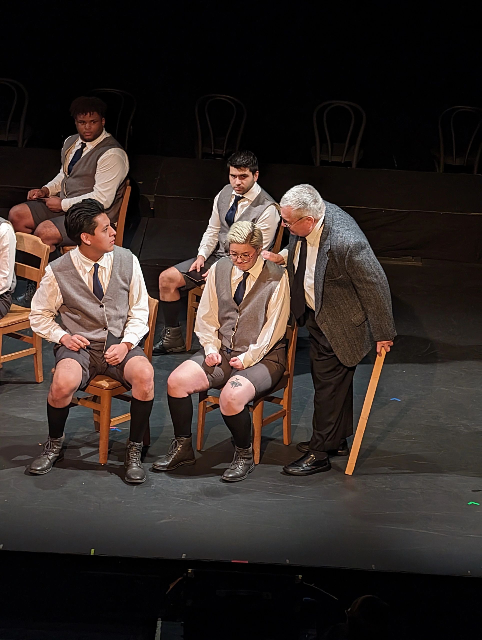 Actors in vests and shorts on stage with an instructor leaning in.