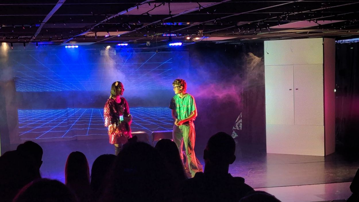 Two actors performing on a stage with neon lights and fog effects.