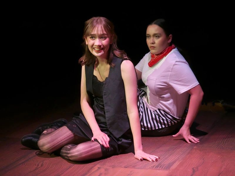 Two women sitting on a wooden floor with contrasting expressions.