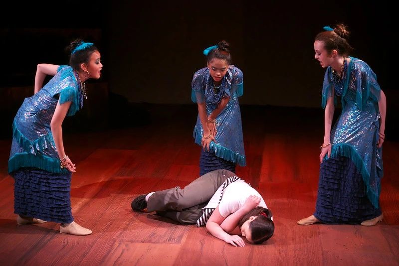 Three women in blue dresses focus on a man lying on the floor.
