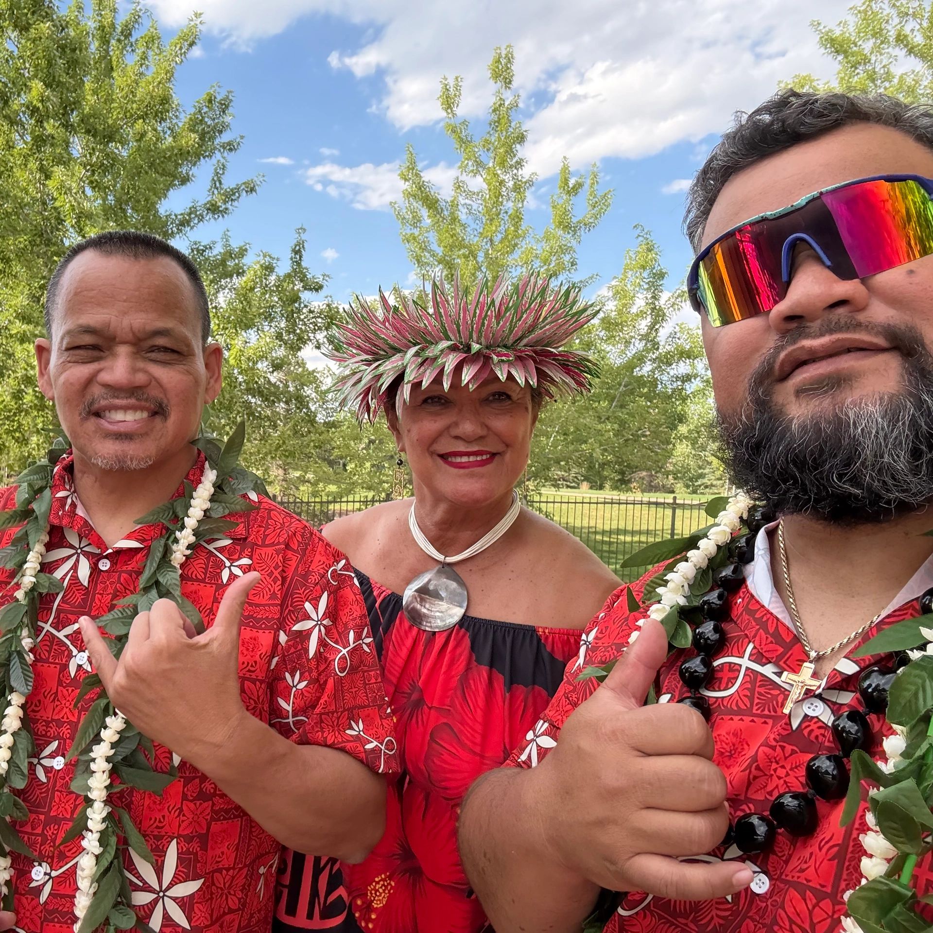 Three people in red Hawaiian attire posing outdoors with hand gestures and a flower crown.