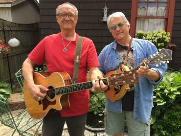 Two older men playing guitar and mandolin outdoors in a garden.