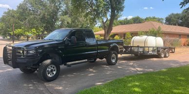 Black pickup truck towing a trailer with large white tanks in a suburban neighborhood.