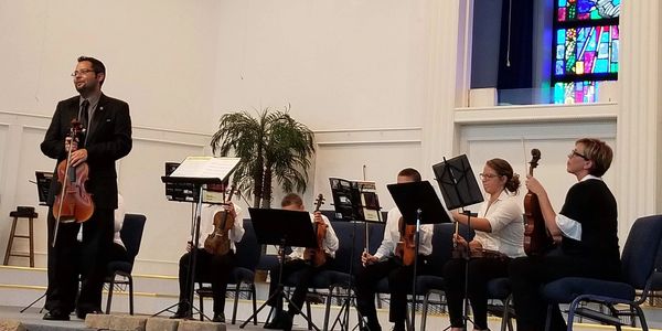 Musicians seated with violins, one standing, in a church setting with stained glass.