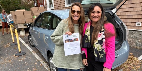 Two women smiling by a car, holding a donation thank-you note.