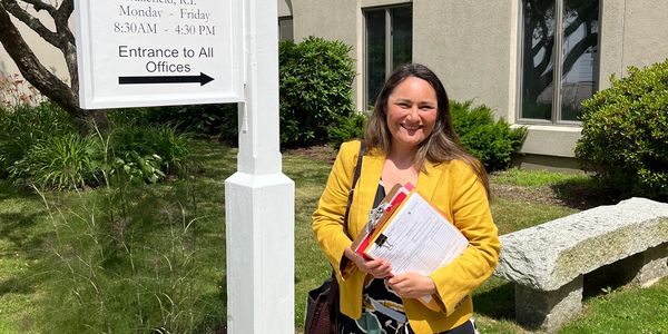 A woman in a yellow jacket stands beside the South Kingstown Town Hall sign in Rhode Island.