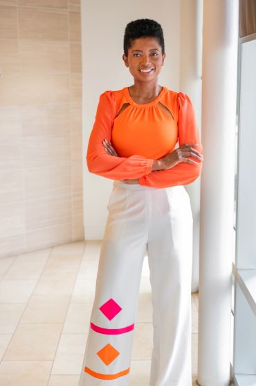Confident woman in an orange top and decorated white pants.