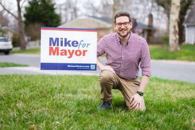 Man kneeling next to a 'Mike for Mayor' campaign sign on a lawn.