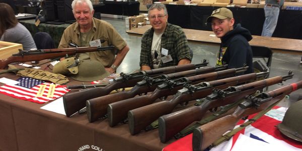 Three men at a table displaying vintage rifles and military helmets.
