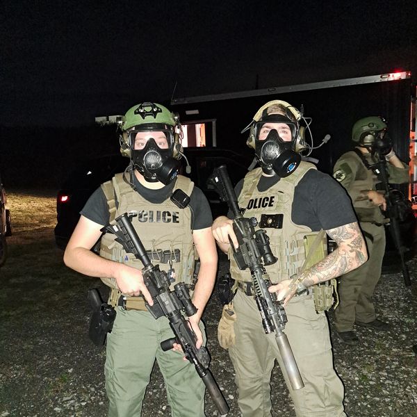 Two police officers in tactical gear and gas masks holding rifles at night.