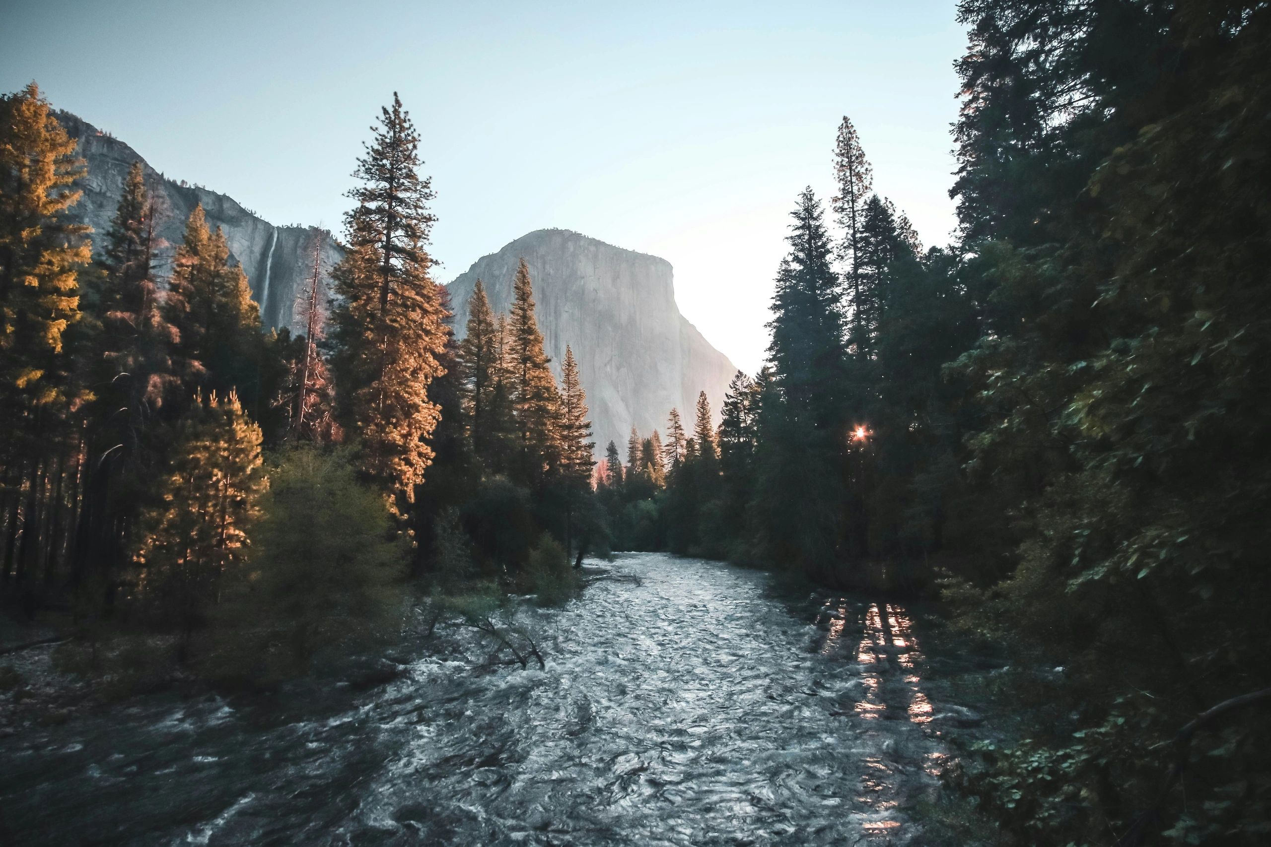 A serene river flows through a forest with mountains in the background at sunset.