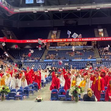 Graduates in red and white gowns throw caps in a celebratory indoor ceremony.