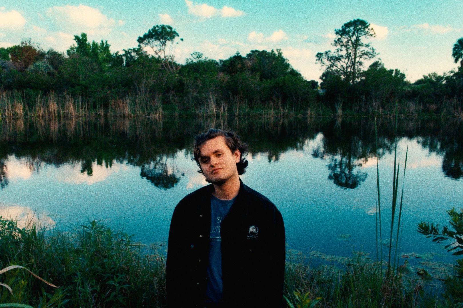 Young man standing near a calm lake with trees reflected on the water.
