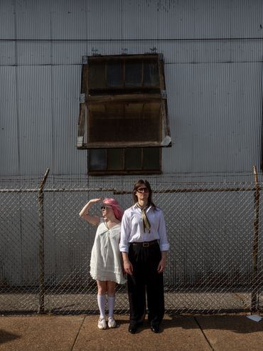 Two people stand against a fence and industrial building.