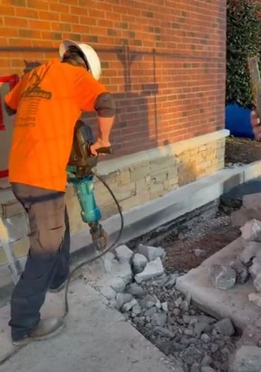 Construction worker using jackhammer on concrete near a brick wall.