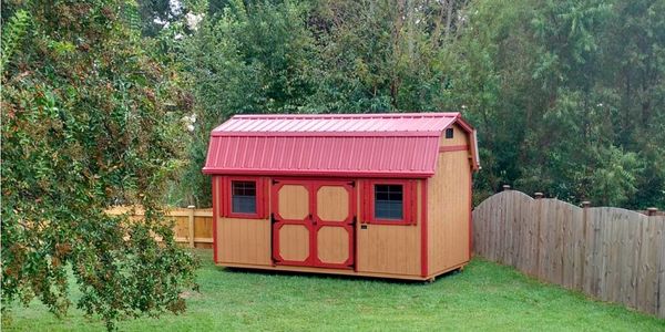 A red and tan garden shed in a fenced backyard surrounded by trees.