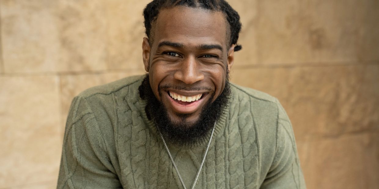A black man who is a therapist in Michigan, with braided hair smiles warmly wearing a green sweater.
