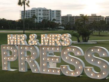 Illuminated "MR &amp; MRS PRESS" sign on a golf course at dusk.