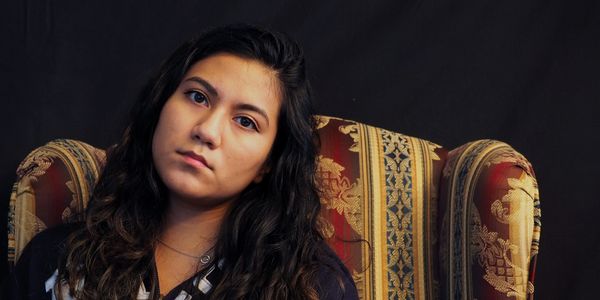 Young woman with long dark hair sitting on a patterned armchair against a dark background.