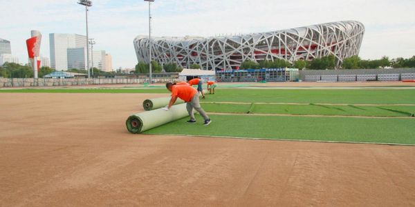 Workers laying artificial turf on a sports field near a large stadium.