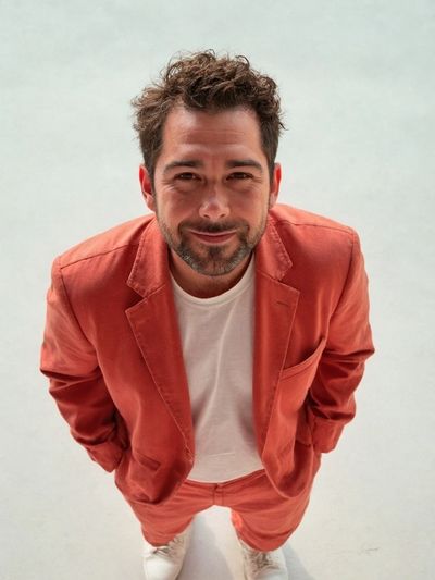Man in a rust-colored suit and white sneakers smiles warmly at the camera.