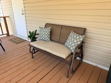 A beige cushioned bench with patterned pillows on a wooden porch.
