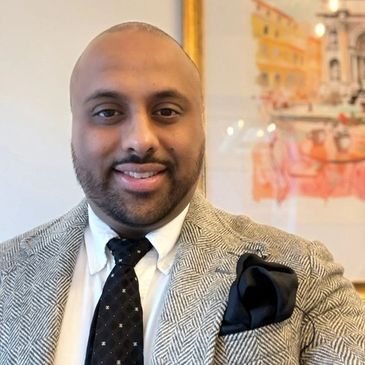 Man in a herringbone suit jacket with a black tie and pocket square smiling.