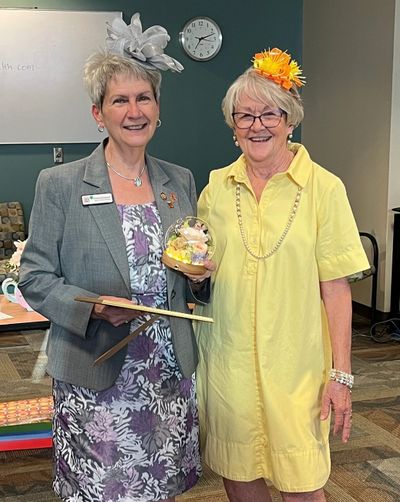 Two smiling older women wearing decorative hats, one holding a plaque and gift.