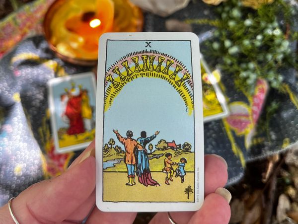 A hand holding the Ten of Cups tarot card with a warm, colorful background.