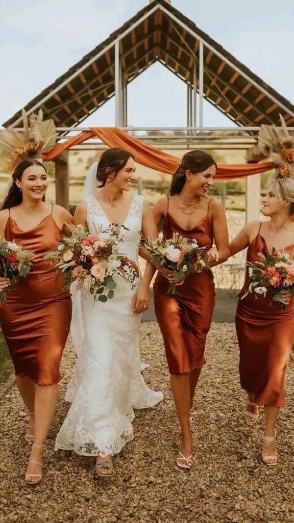 Bride and bridesmaids in satin dresses holding bouquets and smiling outdoors.