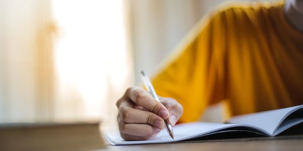 Person writing in a notebook with a pen, wearing a yellow shirt.