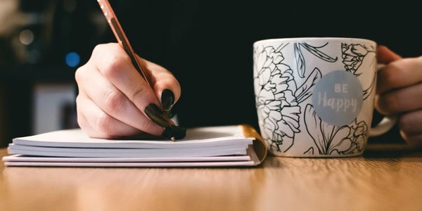 A person writes in a notebook next to a floral mug with 'Be Happy' text.