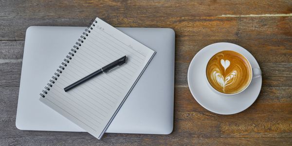 Notebook and pen on a laptop with a coffee cup on a wooden table.